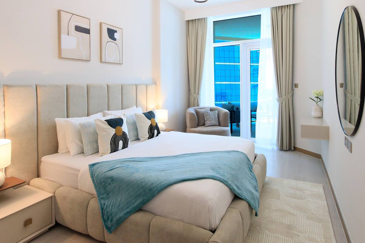 A modern hotel room with a large bed, a nightstand with a lamp, a chair, a balcony with curtains, and two framed artworks on the wall.