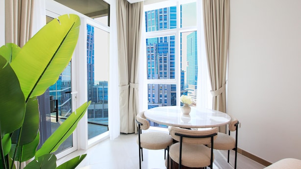 A modern room with a round table and four chairs, large windows with curtains, and a view of tall buildings. - Spacious 1BR Apt Designed by Missoni (Dubai)