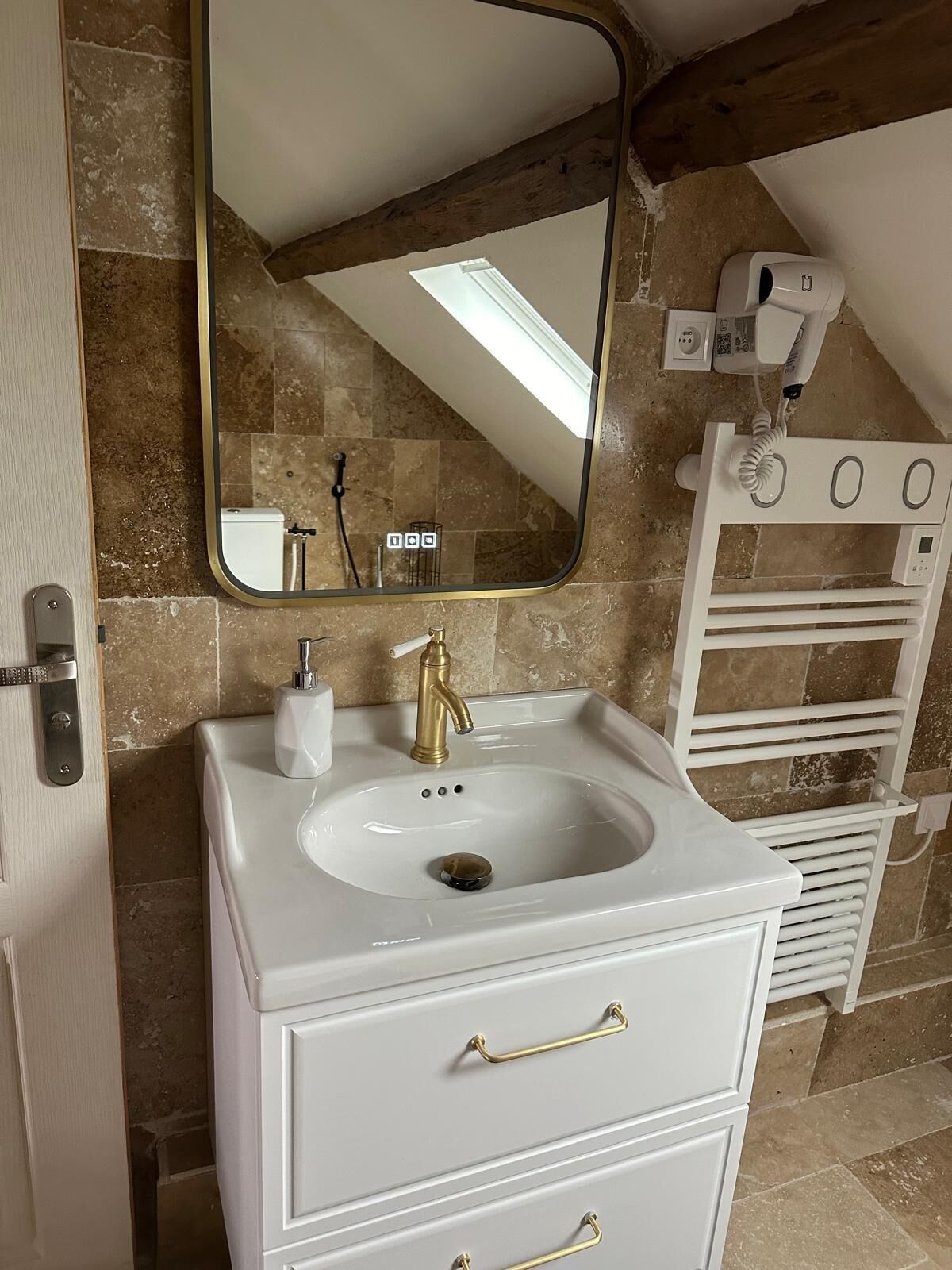 A bathroom with a white sink, gold faucet, and a mirror above it. There is a towel rack to the right and a door on the left.