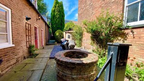A narrow brick alleyway with a circular brick well, a metal trash bin, and a small outdoor seating area with a table and chairs. - Papist Hall Cottages (Barrow upon Humber)