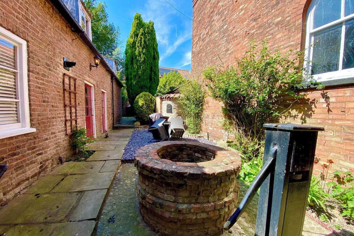 A narrow brick alleyway with a circular brick well, a metal trash bin, and a small outdoor seating area with a table and chairs.