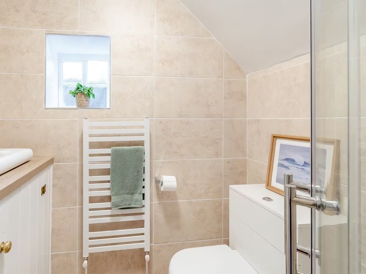 A bathroom with beige tiles, a white toilet, a towel rack with a green towel, a framed picture on the wall, and a window with a plant.