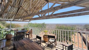 A covered outdoor seating area with wooden tables and chairs, potted plants, and a view of a hilly landscape. - Luxury 2 Story Loft in Boutique Lodge 2 With Free Savannah Adventure 4x4 Tour (Victoria Falls)