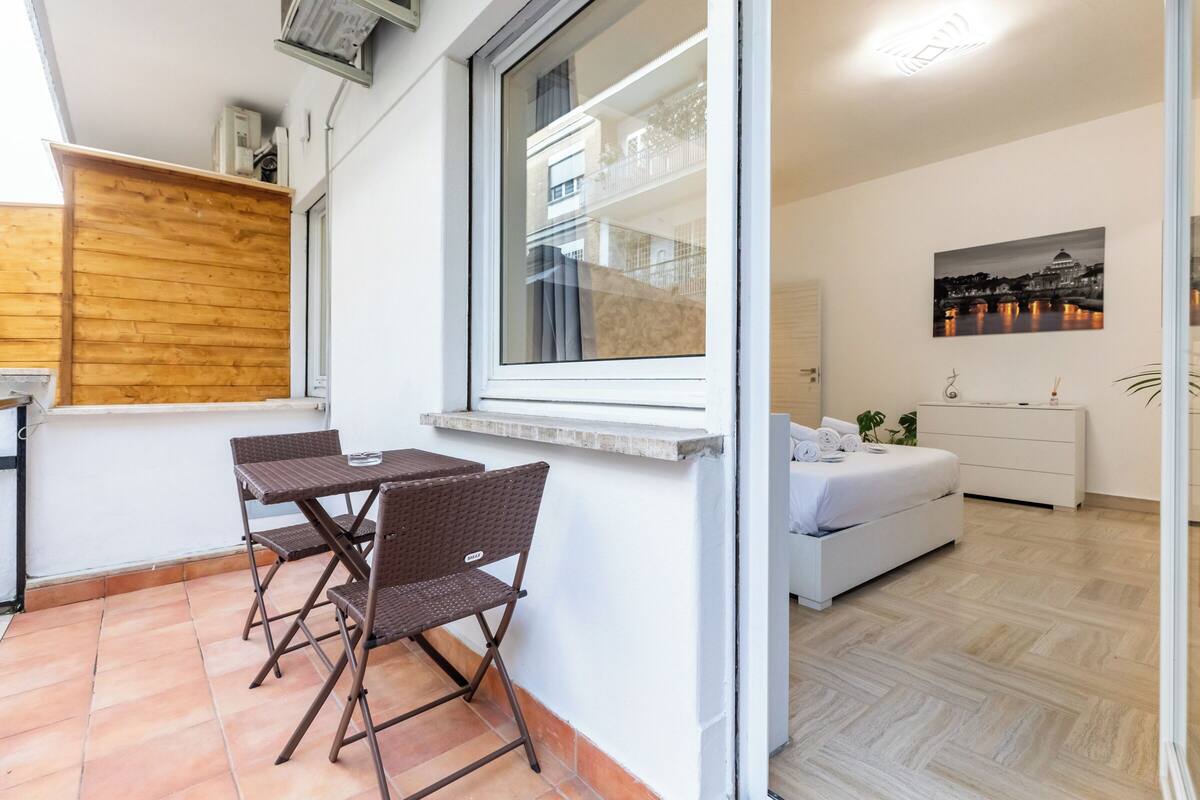 Een gezellig, compact leefruimte met een eethoek, een witte bank, een kleine keuken en een groot raam met uitzicht op een gebouw.