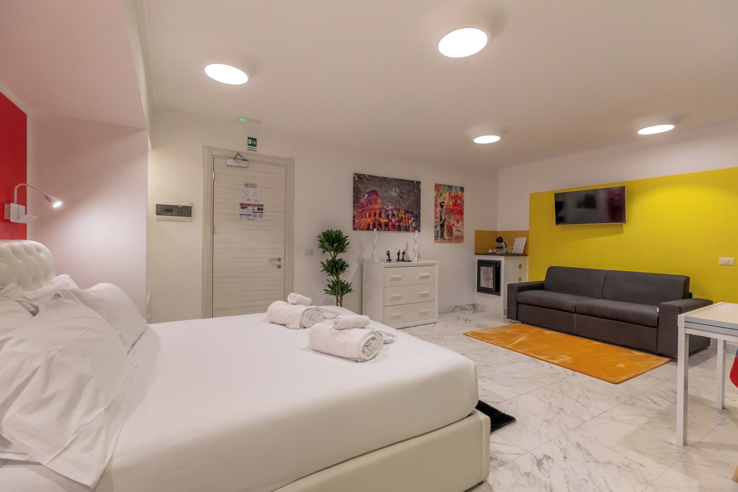 Une chambre d’hôtel moderne avec un grand lit, un canapé gris, une petite table et un téléviseur fixé au mur.