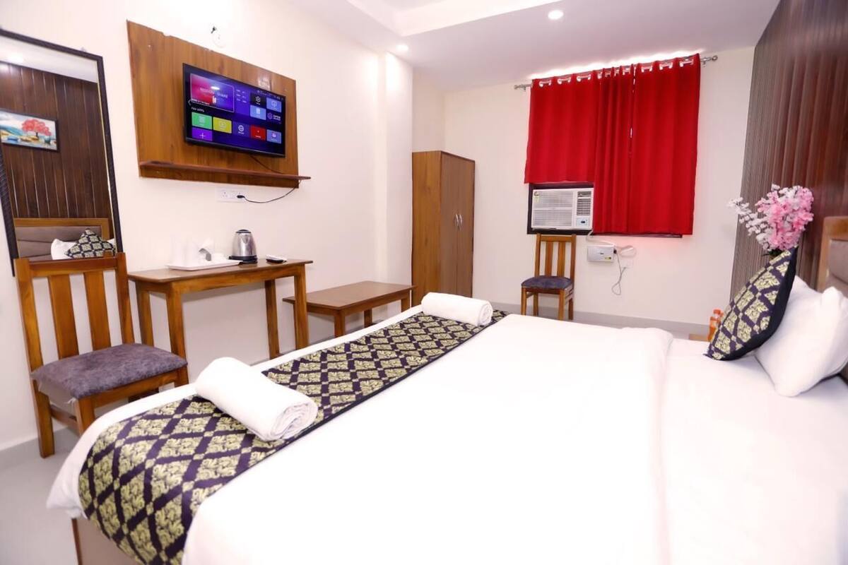 A hotel room with a bed, a TV mounted on the wall, a desk with a chair, a wardrobe, and a window with red curtains.