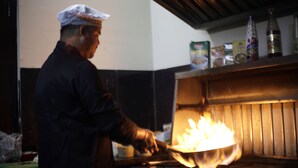 Uno chef cucina con una fiamma visibile sullo sfondo.