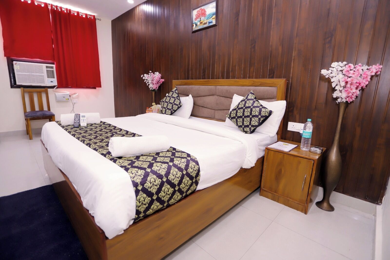 A neatly made bed with patterned bedding, a wooden headboard, and a nightstand with a bottle of water and a vase of flowers.