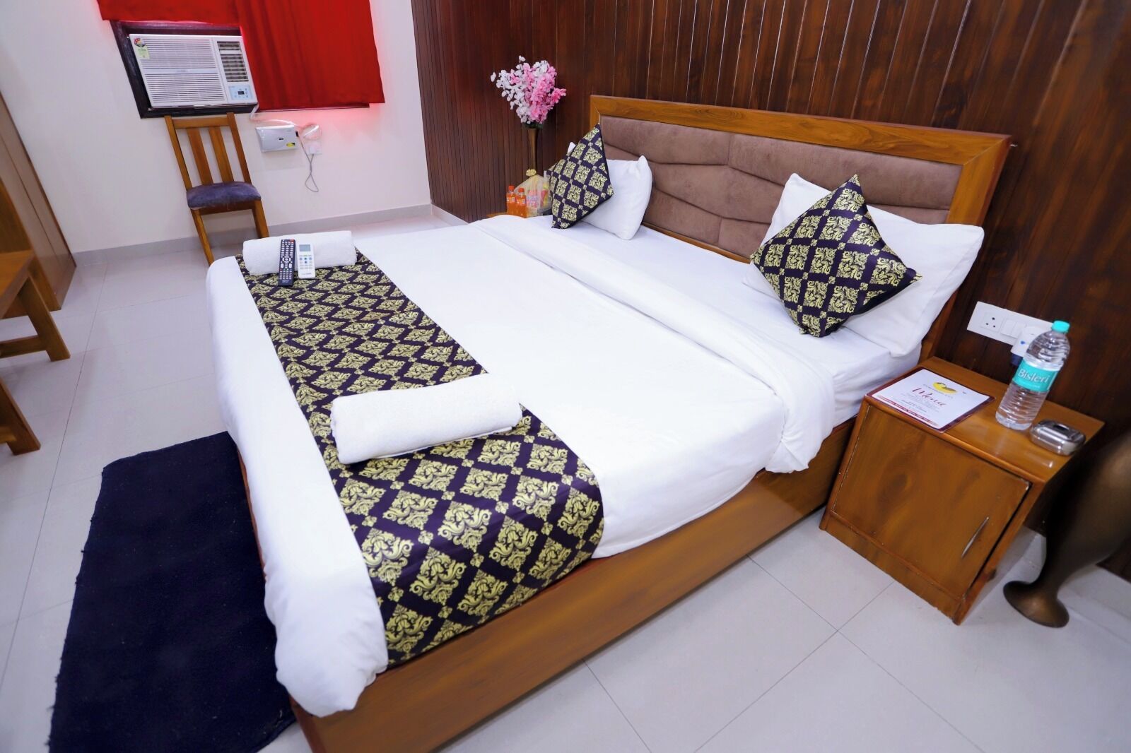 A hotel room with a large bed, a wooden headboard, a bedside table with a water bottle, and a chair.