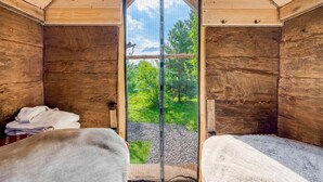A wooden cabin room with two beds, a large window, and a view of greenery outside.