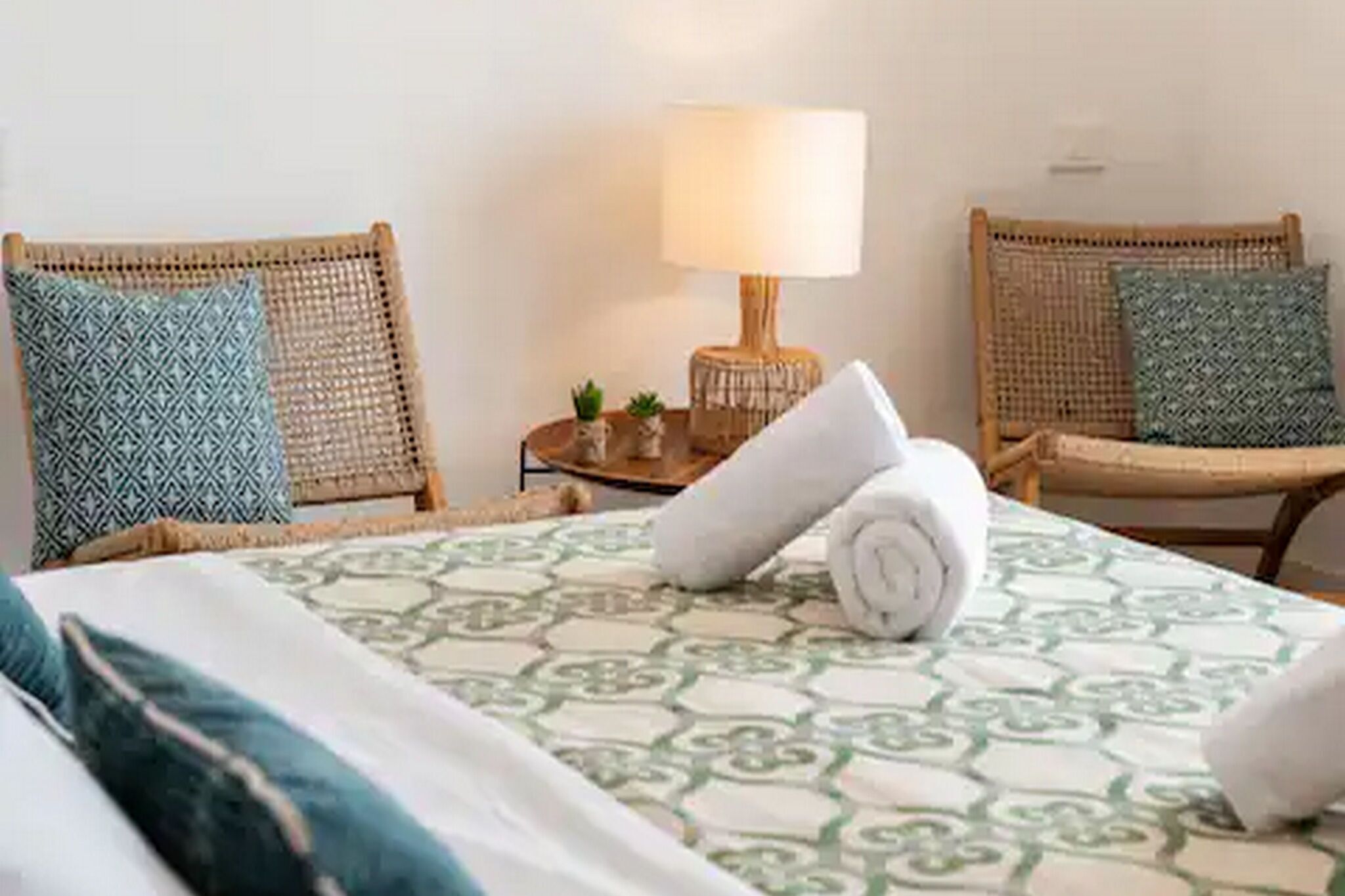 Una camera d'albergo con un letto, due poltrone in vimini, un tavolino con una lampada e cuscini decorativi.