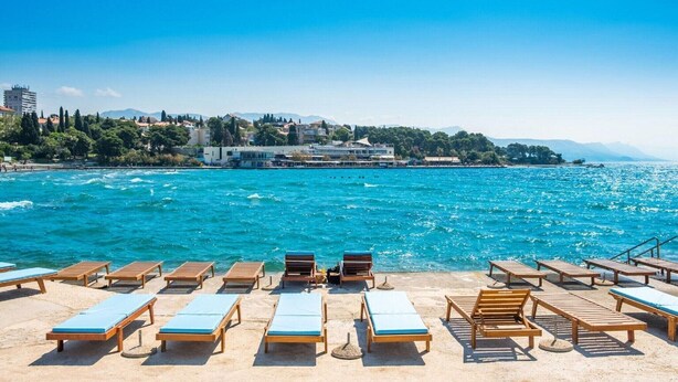 A beach with wooden sun loungers and a clear view of the sea and buildings in the background.