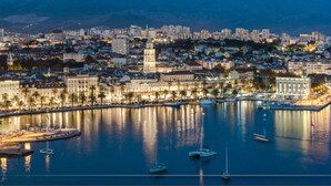 A coastal cityscape at dusk with a prominent waterfront, a central tower, and a dense urban area with buildings and palm trees.