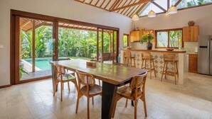 A spacious kitchen with wooden cabinets, a large island with bar stools, and a view of a swimming pool. - Yans Family Villa Canggu by Betterplace (Canggu)