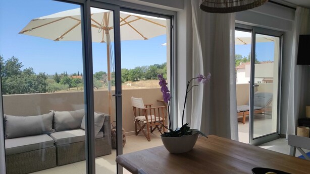 Ein Balkon mit einer Couch und Stühlen, einem Tisch mit Obstschale und Blick auf Bäume und Gebäude.