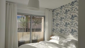 Ein Schlafzimmer mit Bett, weißen Vorhängen, einem großen Fenster mit Blick auf Gebäude und einer Wand mit blau-weißem Muster.
