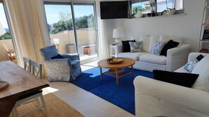 Ein Wohnzimmer mit einem weißen Sofa, einem gemusterten Sessel und einem Holztisch. Ein großes Fenster bietet Blick auf Grünflächen und einen Balkon.