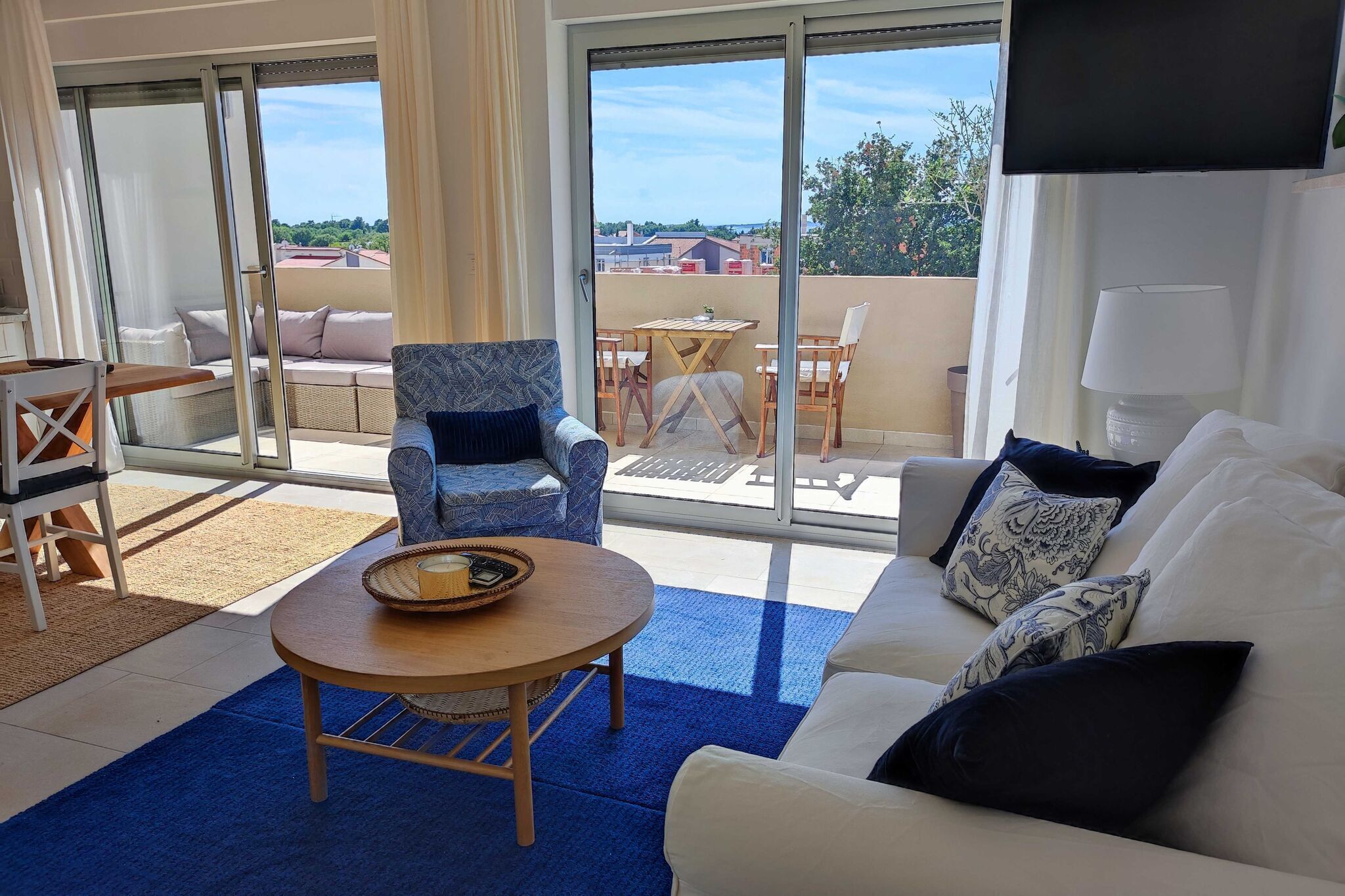A living room with a sofa, armchair, and table, a balcony with a table and chairs, and a TV mounted on the wall.