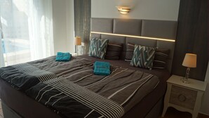Une chambre d’hôtel moderne avec un grand lit, une tête de lit dotée d’un éclairage intégré, deux tables de chevet avec des lampes et une vue sur l’extérieur par la fenêtre.