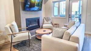 A modern living room with a fireplace, a large flat-screen TV mounted above it, a sofa, two armchairs, and a coffee table. - Nordix Townhome / Private hot tub / New build (Panorama)