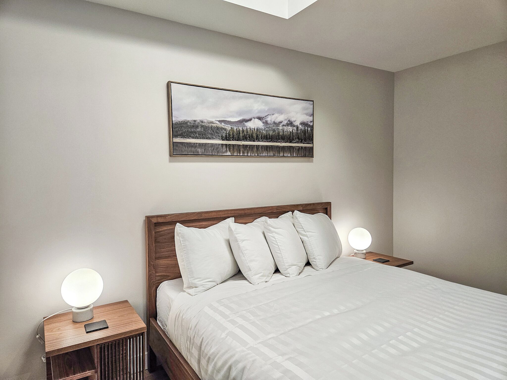 Une chambre à coucher bien aménagée, avec une tête de lit en bois, un grand lit recouvert de linge de lit blanc, deux tables de chevet surmontées de lampes rondes et un tableau représentant un paysage, accroché au mur.