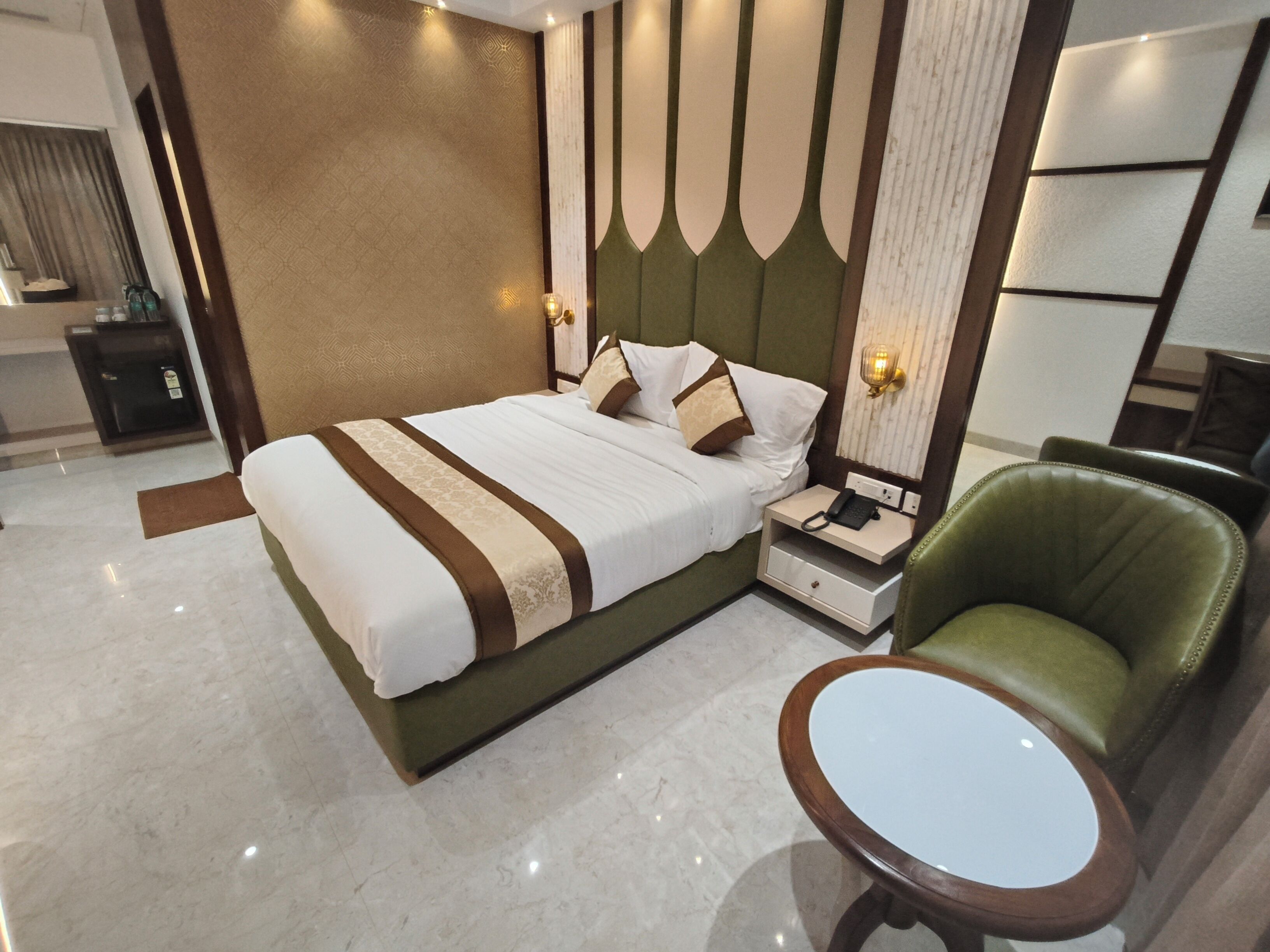 A modern hotel room with a large bed, a bedside table with a phone, a green armchair, and a small round table.