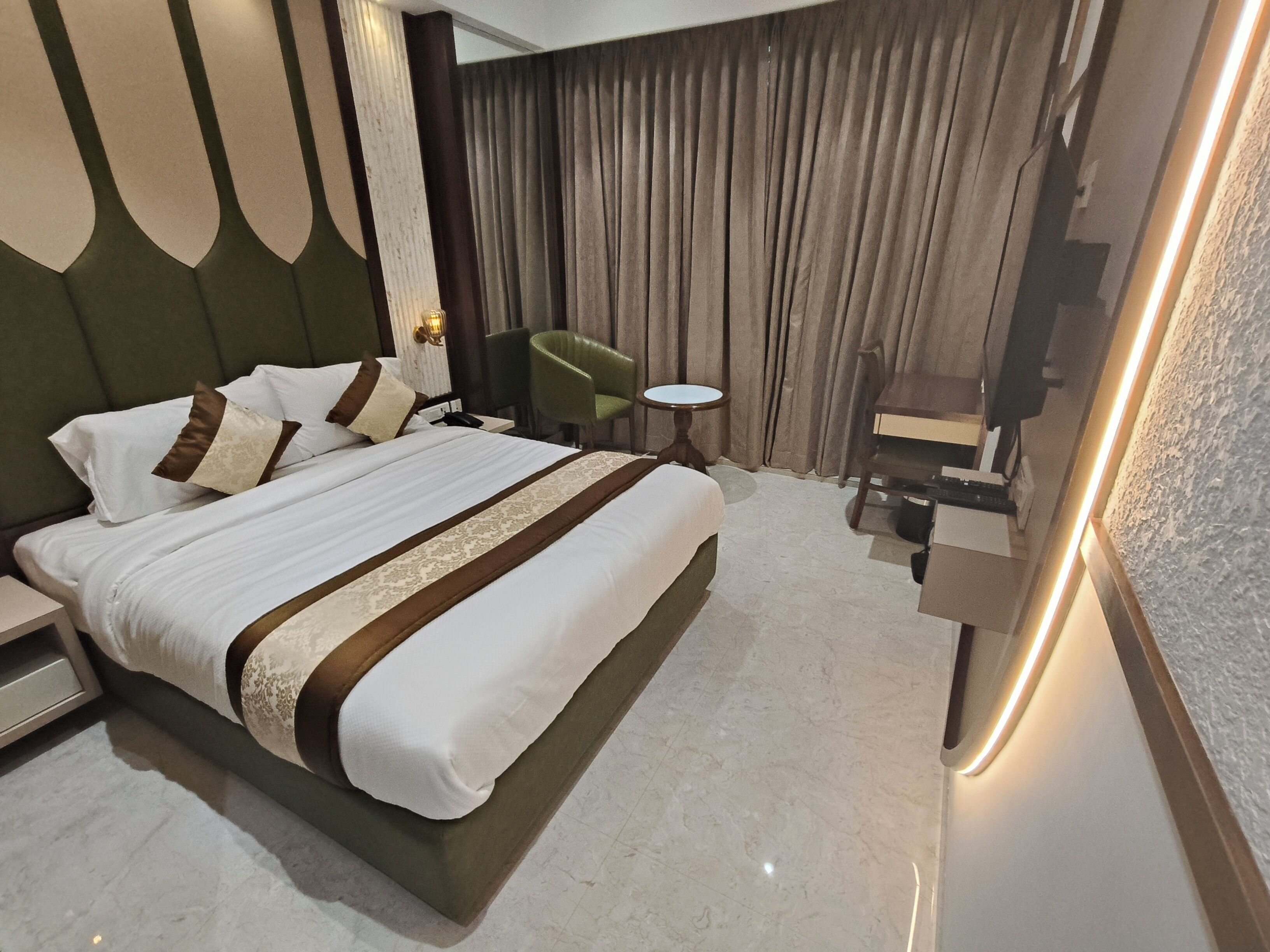 A modern hotel room with a large bed, a small round table, a green chair, and a mirror with a light strip.