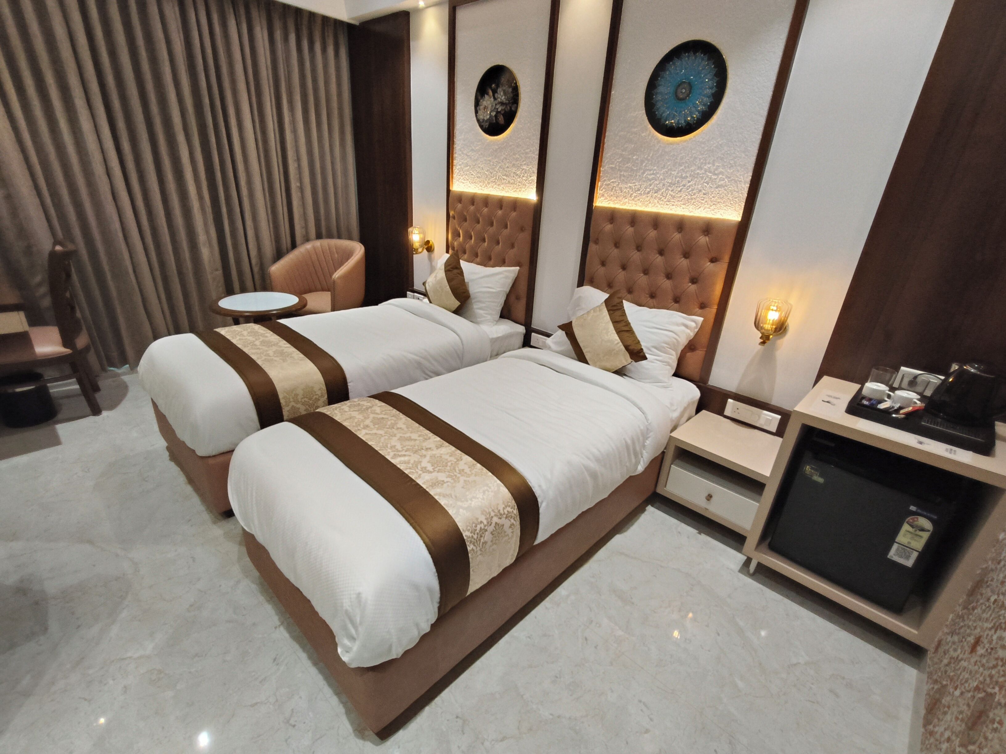 A hotel room with two beds, a small round table, a chair, a minibar, and decorative wall panels.