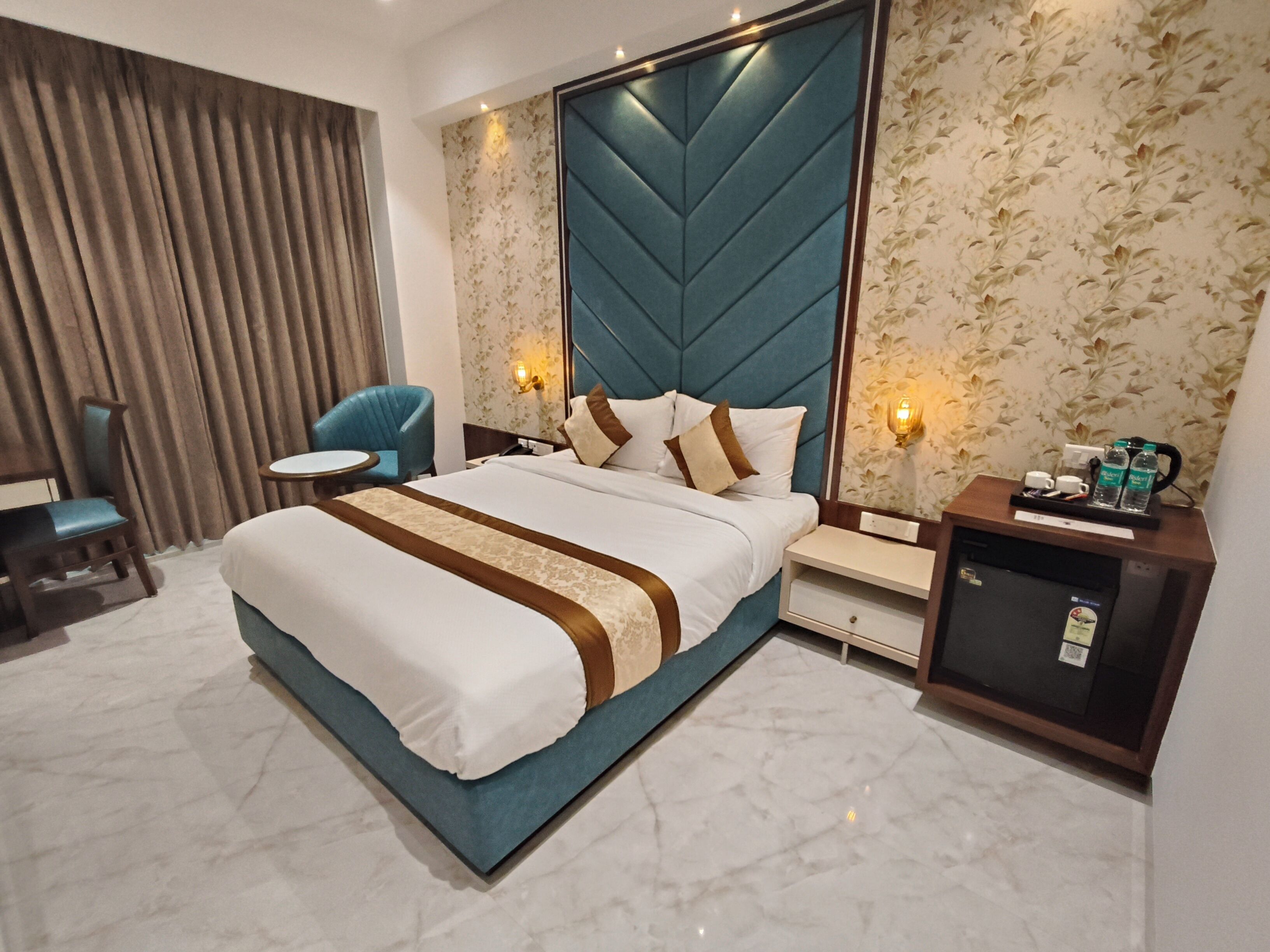 A hotel room with a large bed, a desk, a chair, a minibar, and a wall with floral wallpaper.