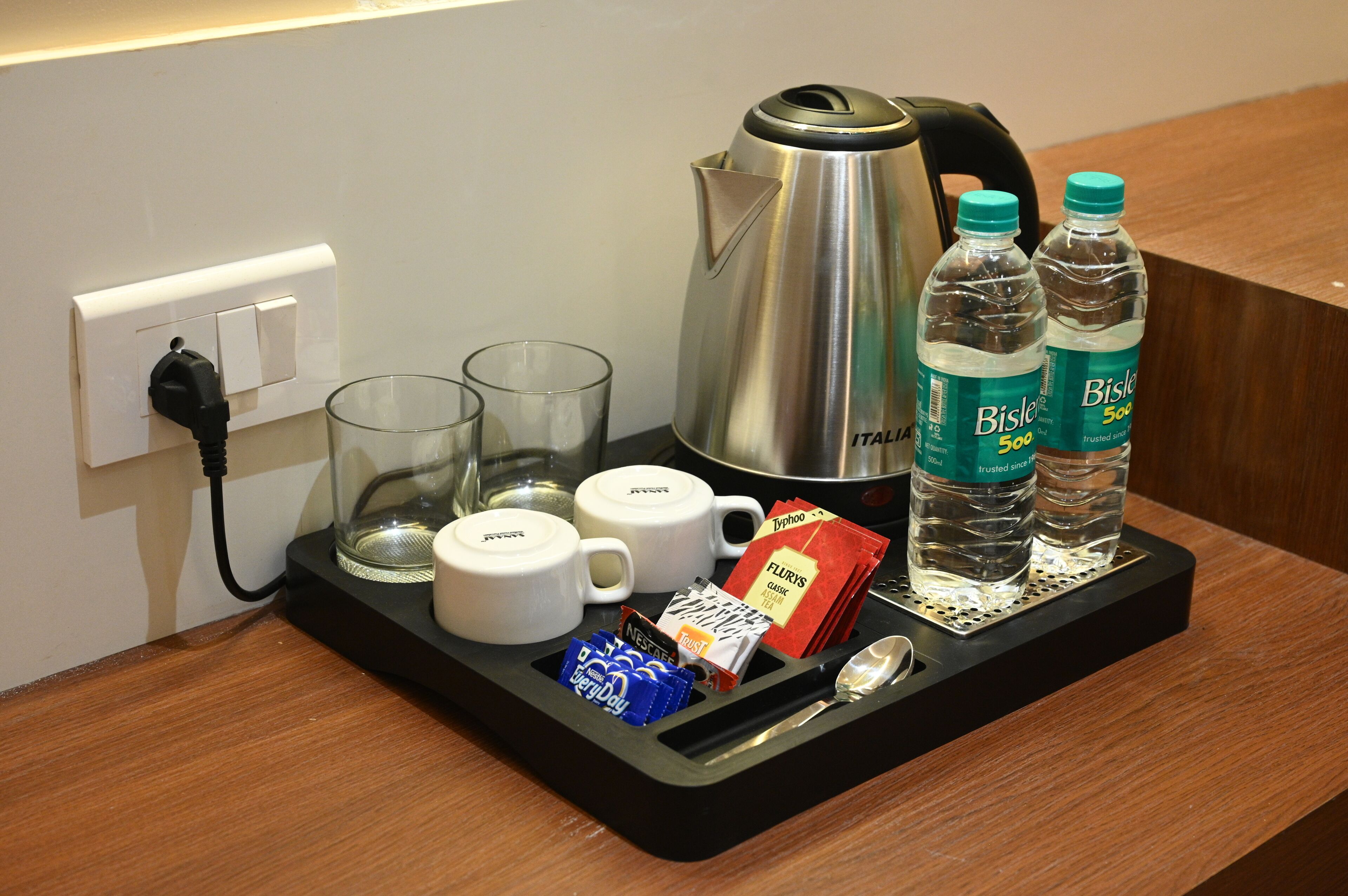 A hotel room's breakfast tray with a kettle, two water bottles, two glasses, a coffee maker, a spoon, and some packaged food items.