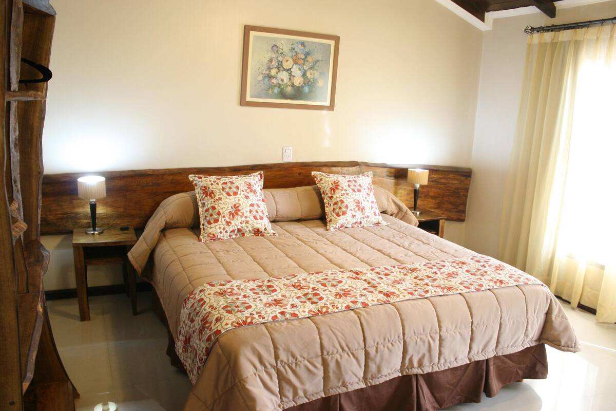 A bedroom with a large bed, two bedside tables, a nightstand, a lamp, and a painting on the wall.