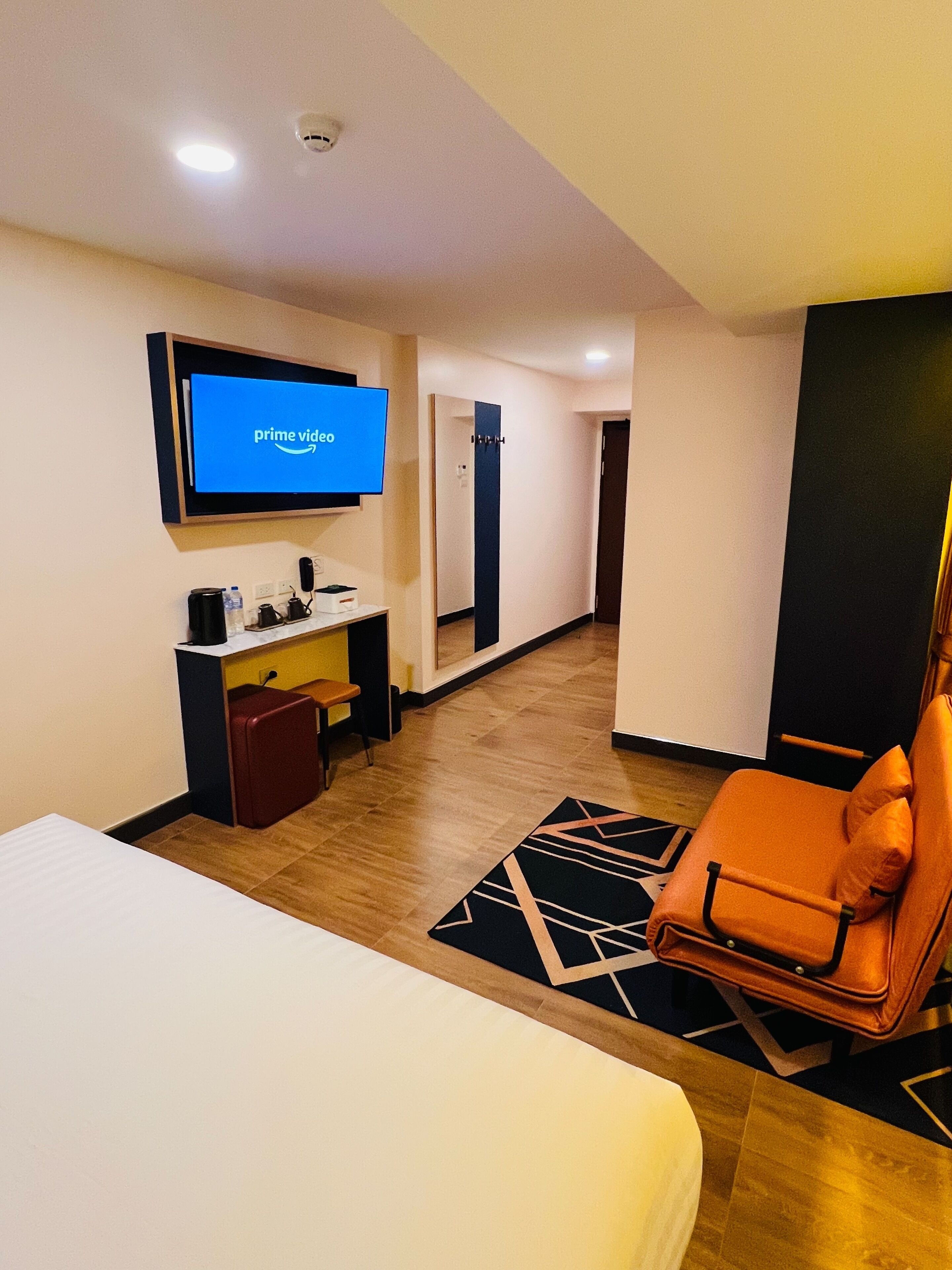 A hotel room with a bed, a desk with a computer, a TV displaying 'prime video', a chair, and a small table with a water bottle and a cup.