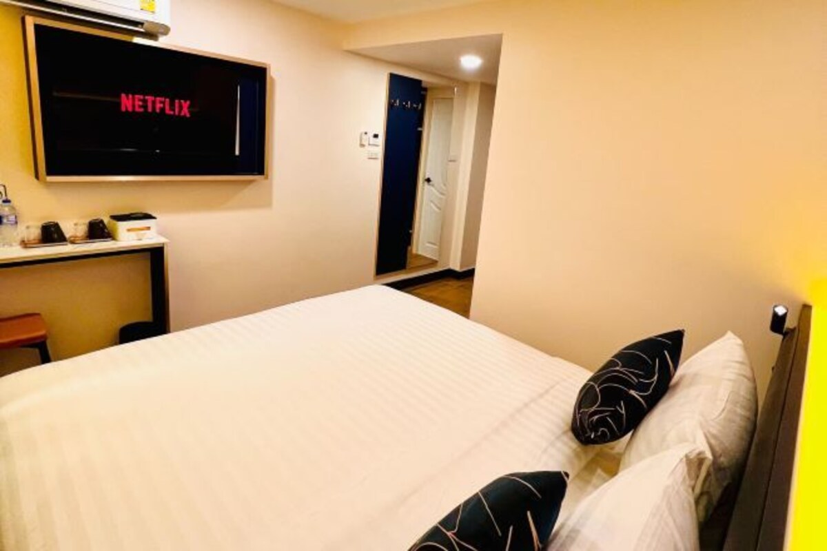 A hotel room with a bed, a desk with a television displaying 'NETFLIX', and a door leading to another room.