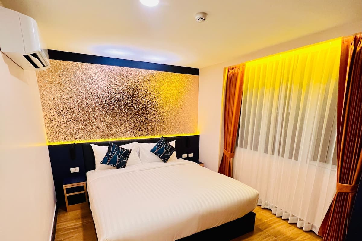 A hotel room with a large bed, wooden flooring, a headboard with a textured wall, and a window with curtains.