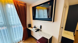 Habitación de hotel con un ventanal grande, un televisor de pantalla plana en la pared, un pequeño escritorio con una cafetera y una maleta roja en el suelo.