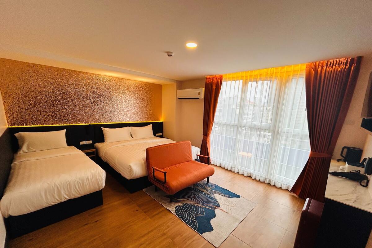A hotel room with two beds, a sofa, a patterned rug, and a view of the city through large windows.