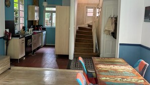 Una cocina con armarios de madera, una mesa de comedor con sillas coloridas y una escalera que sube a otra habitación.