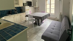 Eine kleine Küche mit weißen Schränken, einem Esstisch mit grauem Tischläufer und einem grauen Sofa.