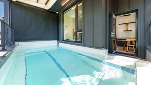 Une piscine à l’eau claire, une terrasse carrelée et un bâtiment moderne doté de grandes fenêtres et d’une porte coulissante donnant sur un espace cuisine.