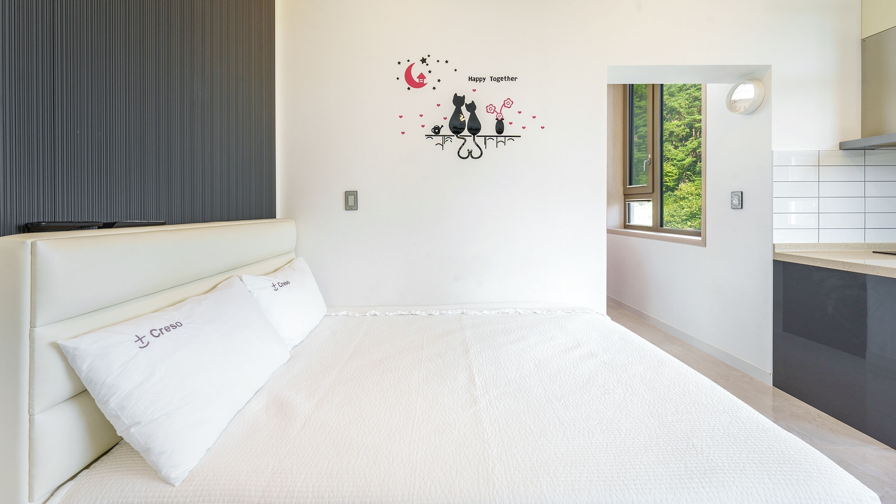 A bedroom with a bed, a nightstand, a wall decal, a window, and a view of greenery outside.