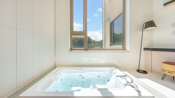 Un baño moderno con una bañera grande, una ventana con vista a follaje y una lámpara de pie.