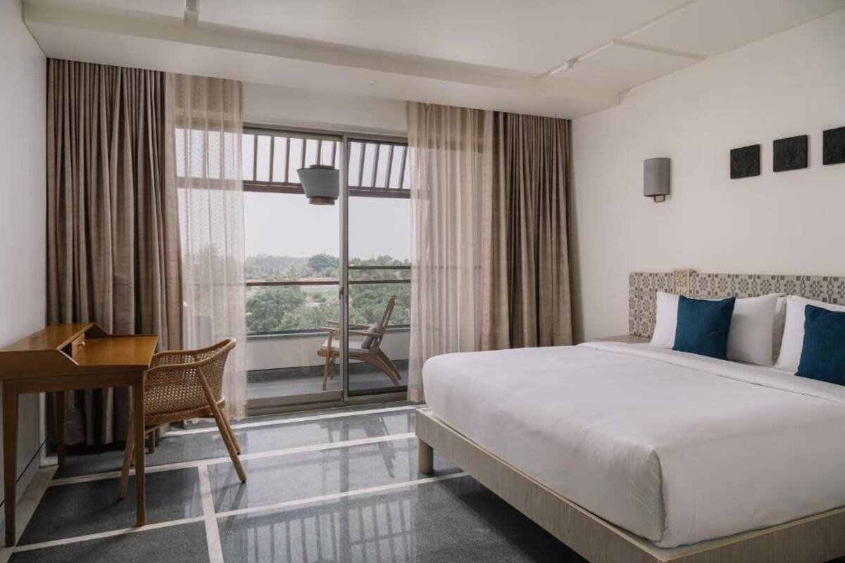 A hotel room with a bed, a desk, a chair, a window with curtains, and a view of greenery.