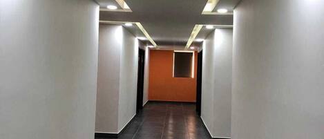 A long, narrow hallway with dark tiled flooring and white walls, leading to an orange door at the end.
