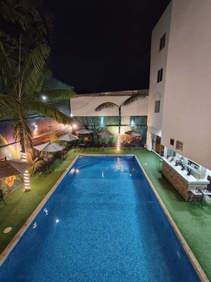 Piscina con sillones de descanso y sombrillas, rodeada por un edificio con un mural.