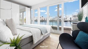 A modern bedroom with a large bed, a view of the water, and a balcony with a chair. - Biscaya - Sauna, Shower, 360° Panoramic Rooftop Terrace, Floating House (Olpenitz)