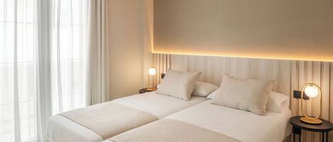 A hotel room with two beds, a large window with sheer curtains, and a wall-mounted light fixture.