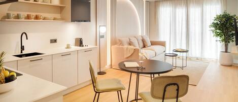 A modern kitchen with white cabinets, a black faucet, and a bowl of fruit. There is a dining area with two chairs and a small table.