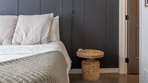 Un dormitorio con cama, una lámpara de noche, una mesita redonda y una puerta que da a otra habitación.