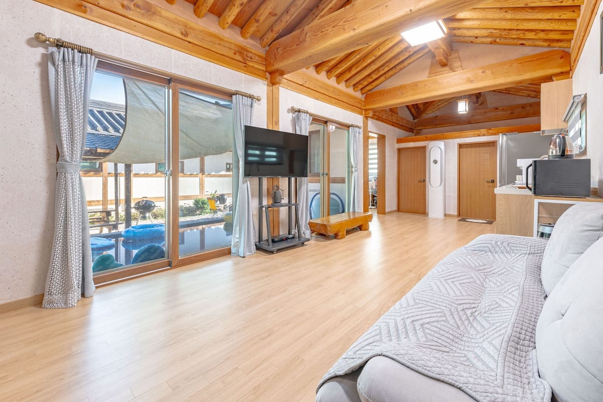 A spacious room with a wooden ceiling, a large window with curtains, a flat-screen TV, a bench, and a sofa.