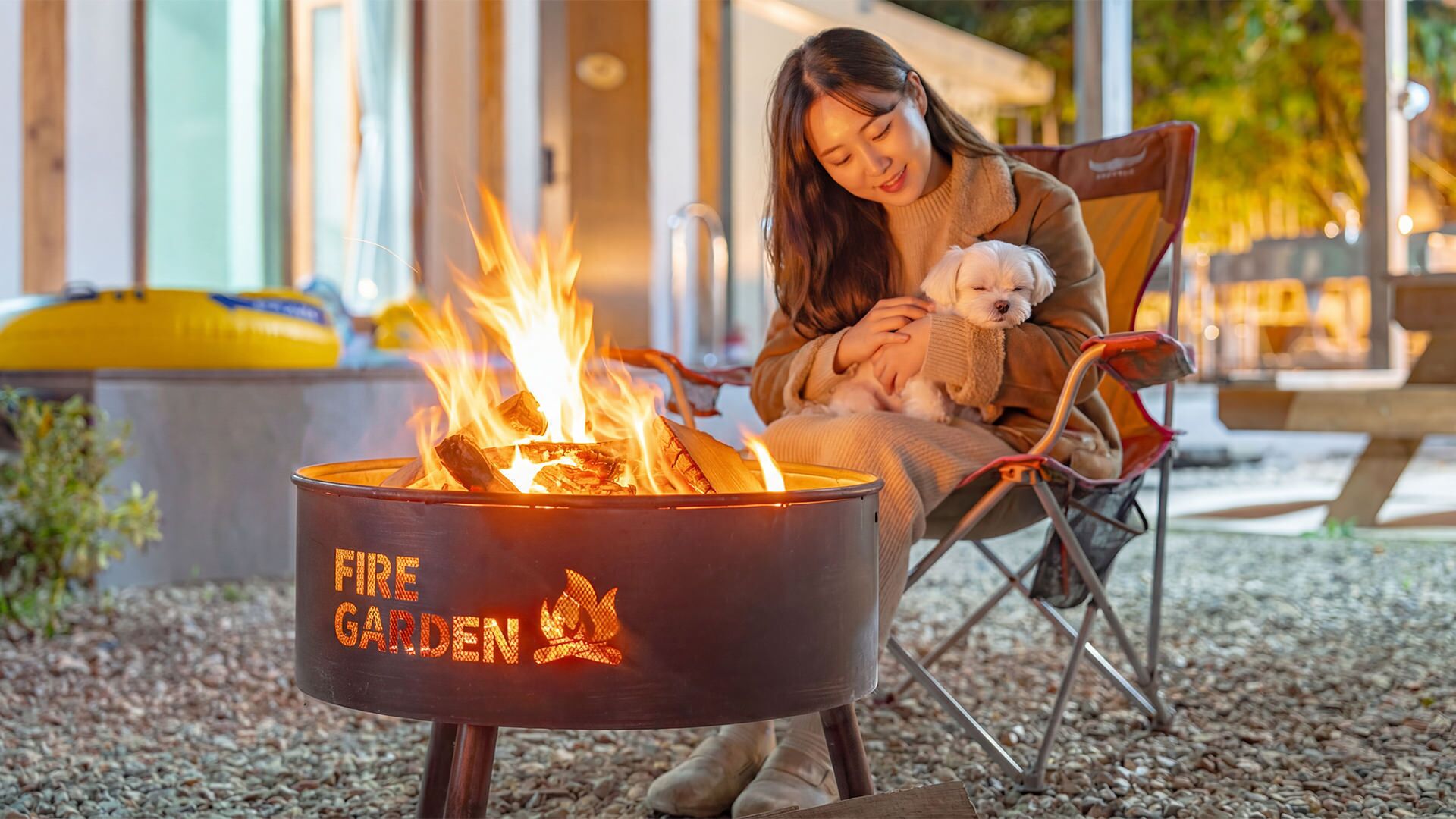 Eine Person sitzt am Feuerkorb, neben einem Hund, im Freien mit einem Gebäude im Hintergrund.